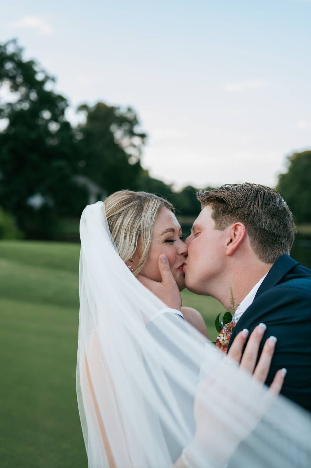 A bride and groom share a romantic kiss outdoors on a green lawn, with the bride wearing a flowing white veil.