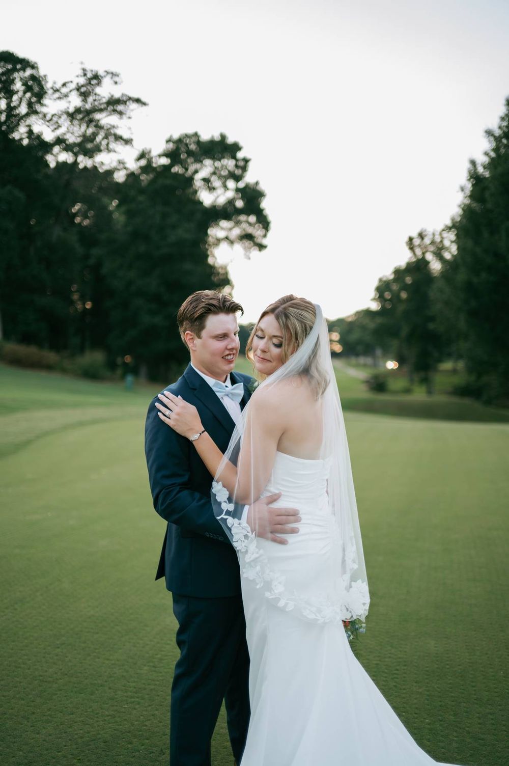 Bride and groom embrace on a golf course at dusk, she wears a white gown with veil, he wears a navy suit.