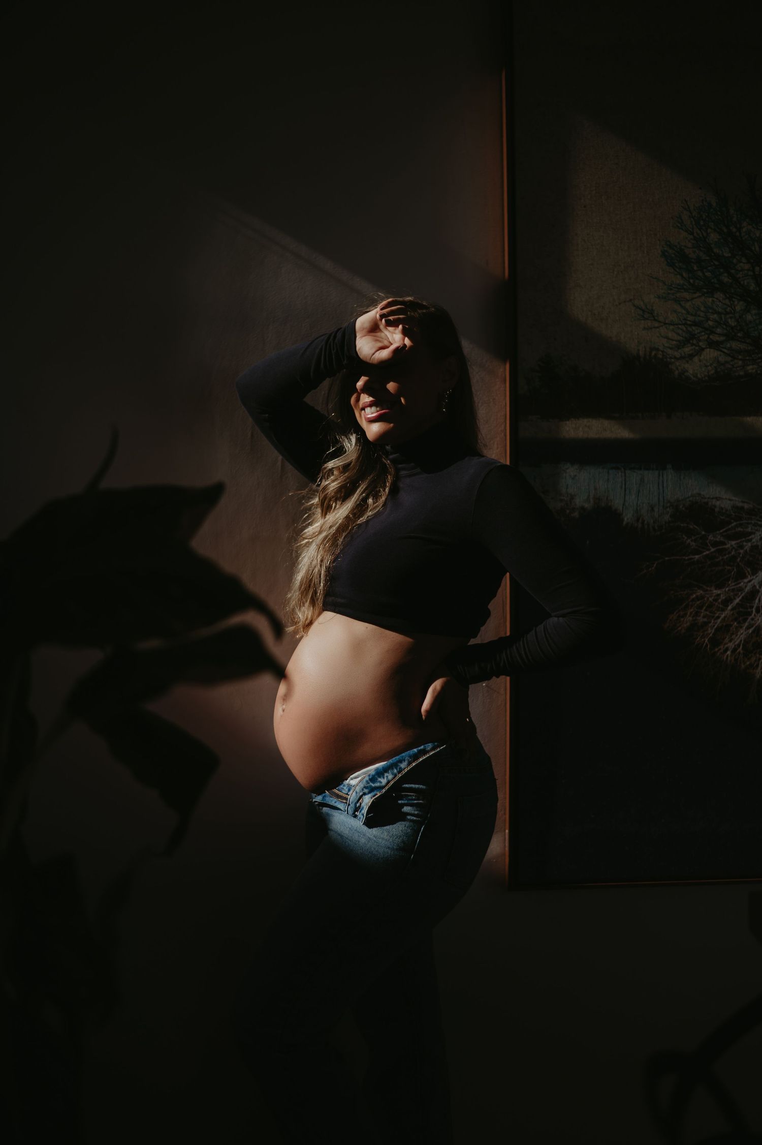 Pregnant woman in crop top and jeans poses in dramatic moody lighting, cradling baby bump against dark wall.