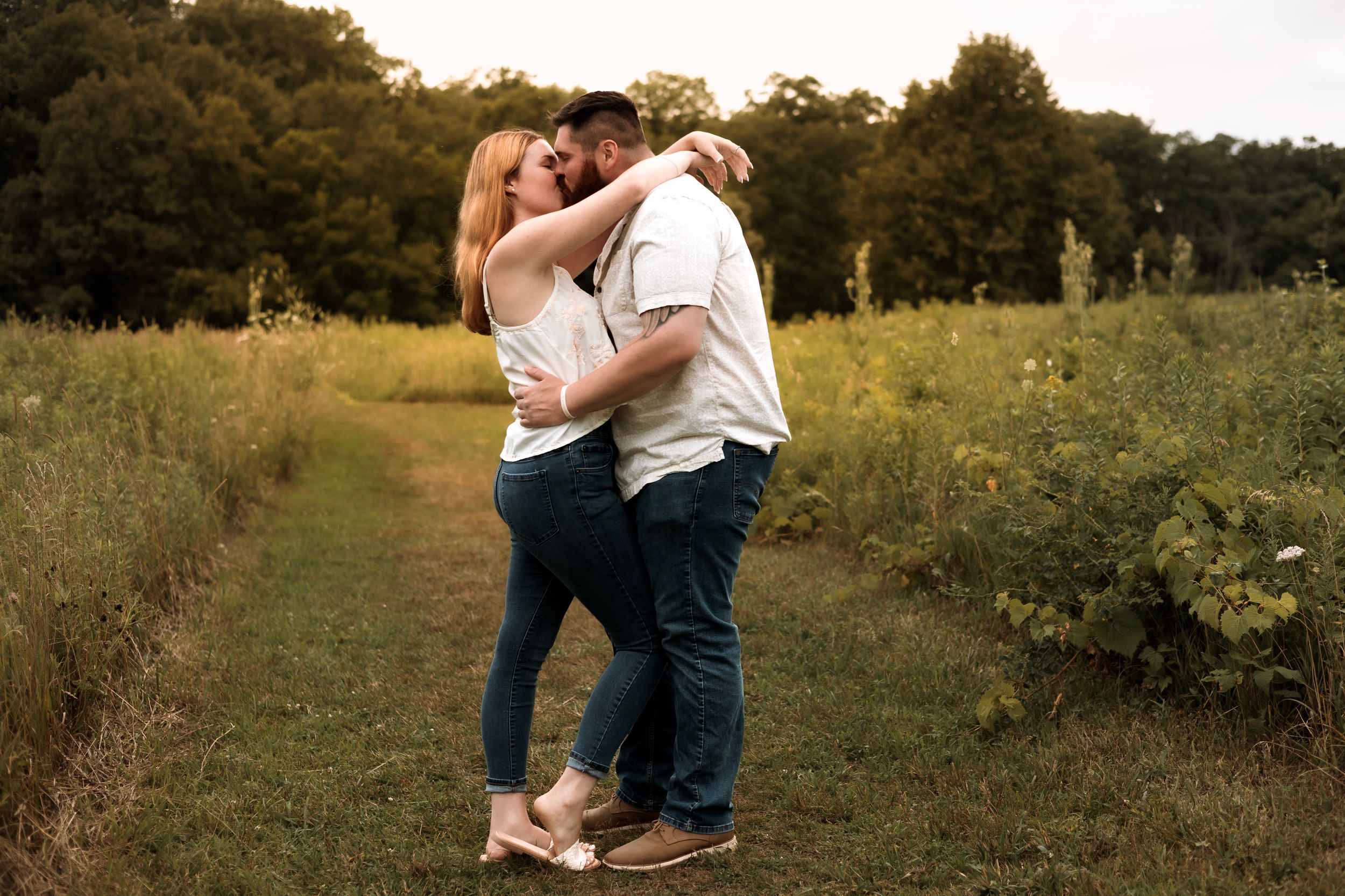 Couple embracing and kissing on a grassy field path surrounded by lush green trees during golden hour.