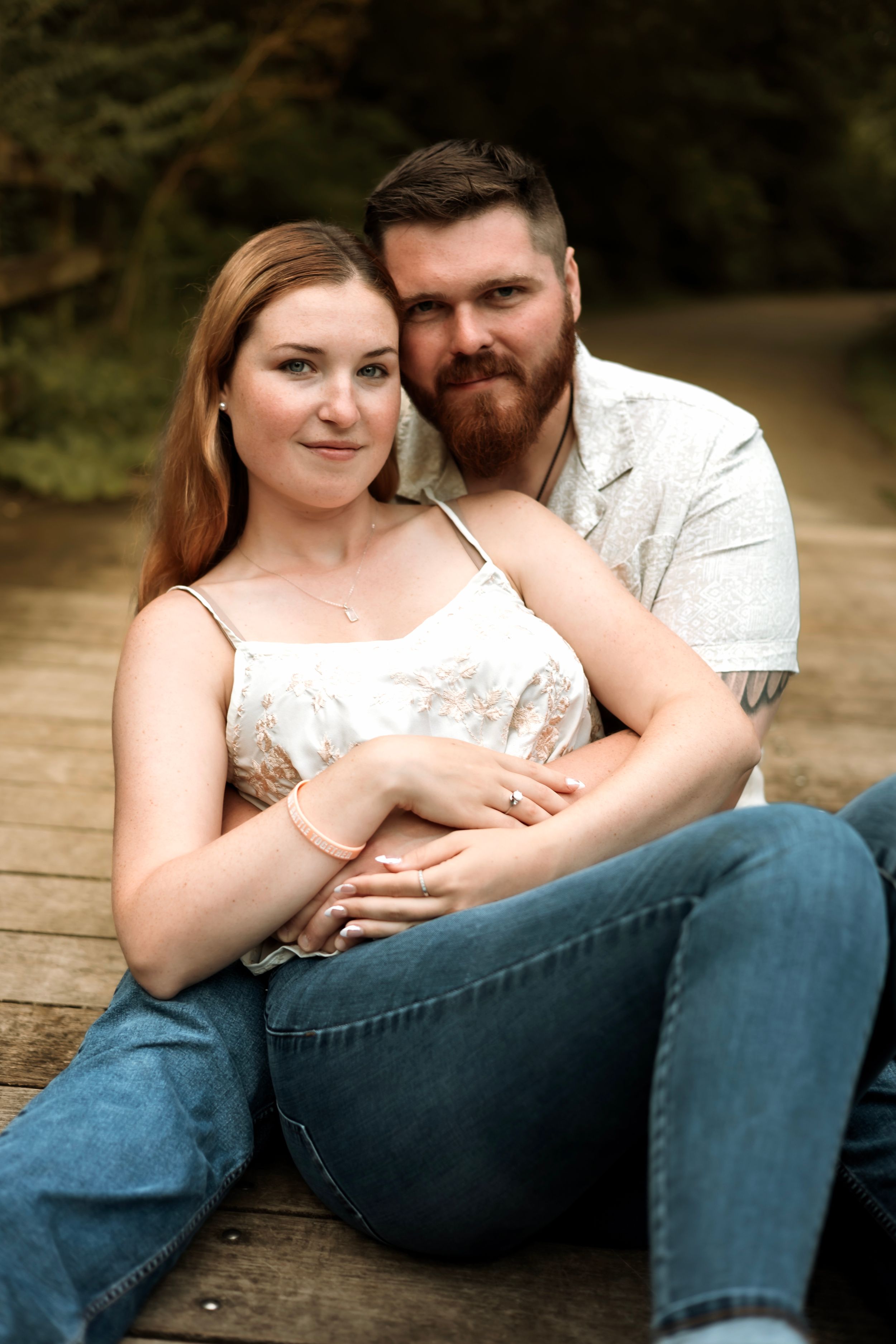 A couple sits together on a wooden deck outdoors, the woman in a floral top and jeans, the man embracing her from behind.