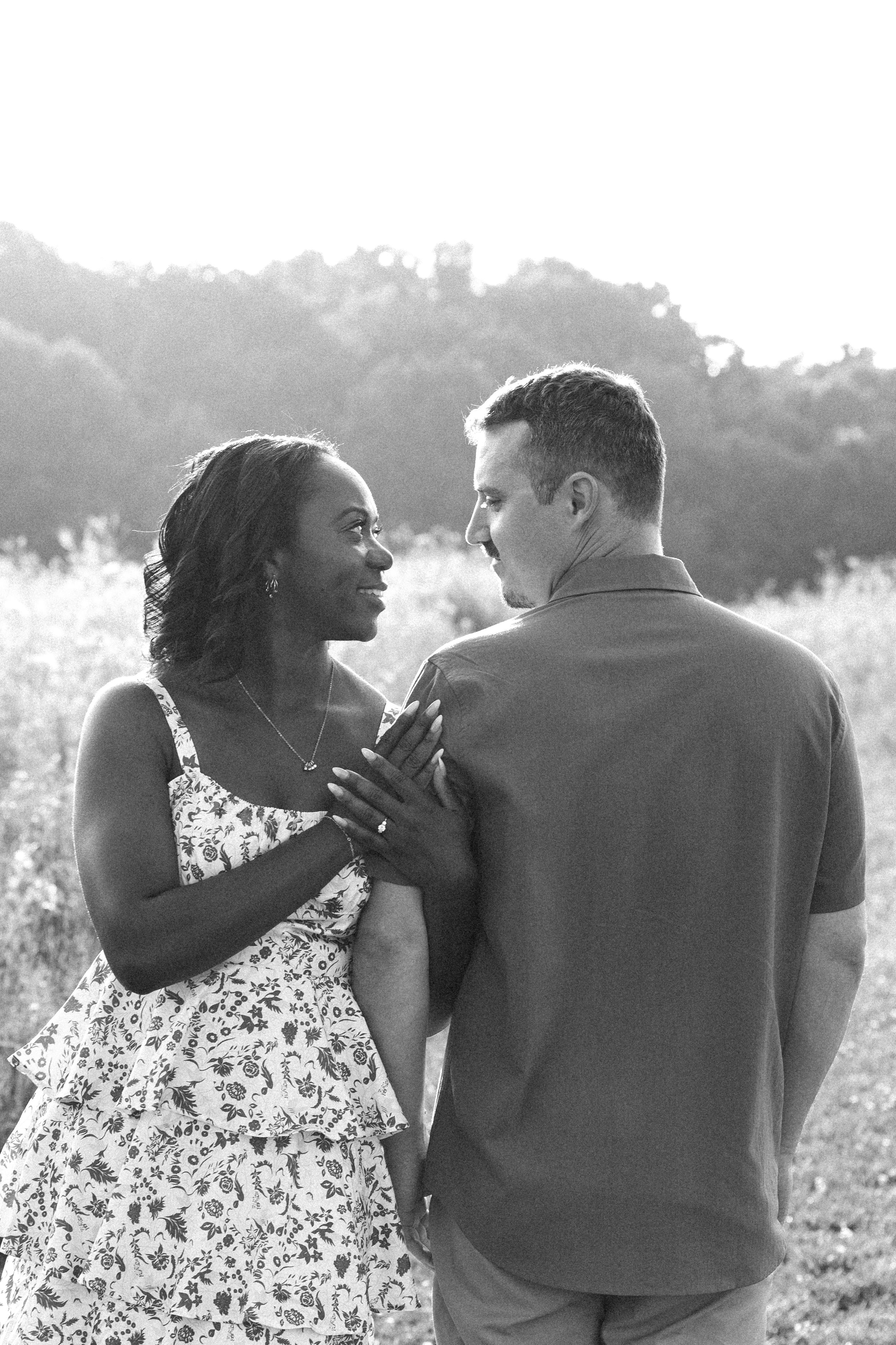 Black and white photo of a smiling couple sharing a tender moment outdoors in a sunlit field.
