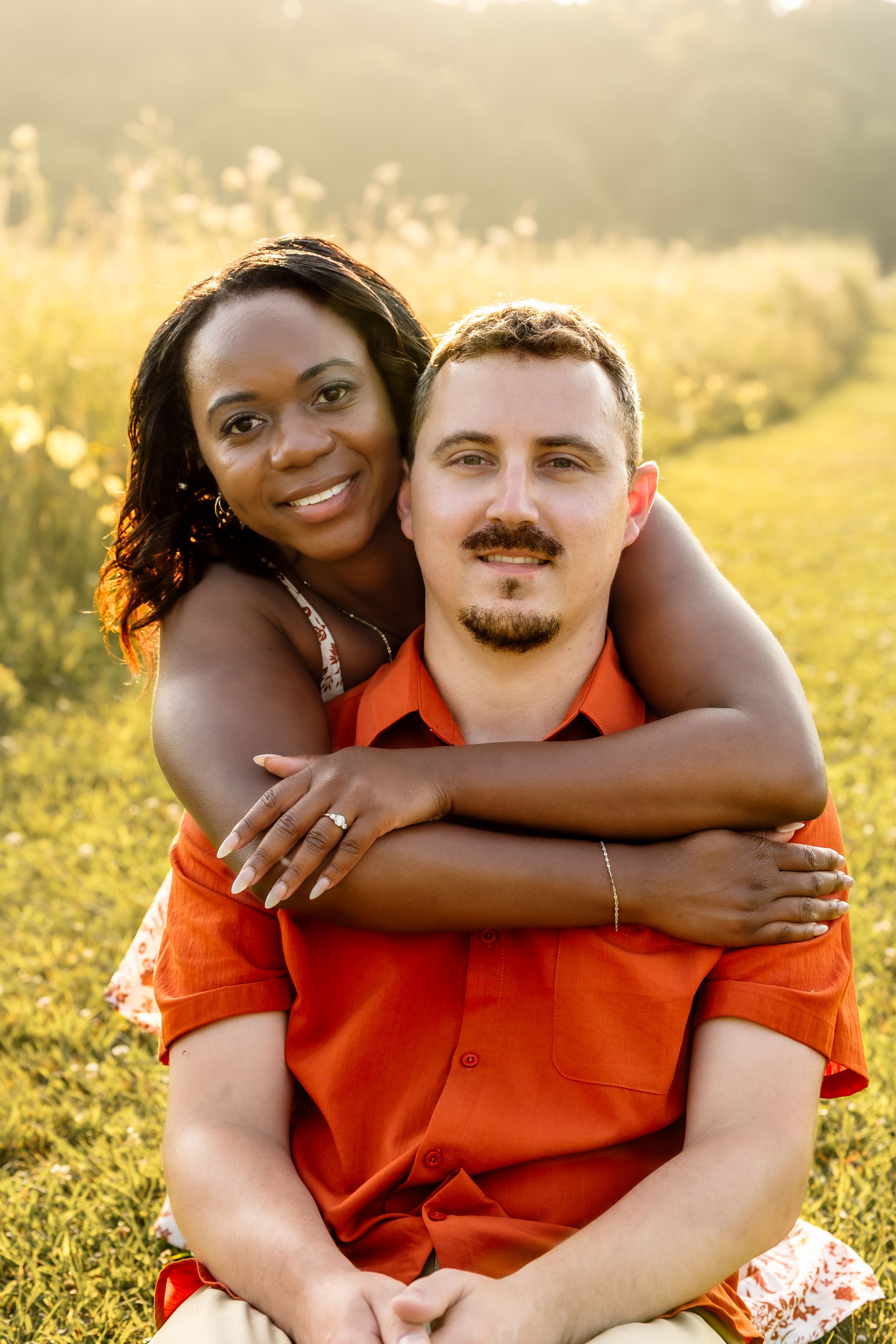 Interracial couple embracing outdoors in a sunny field, woman hugging man from behind, both smiling warmly.