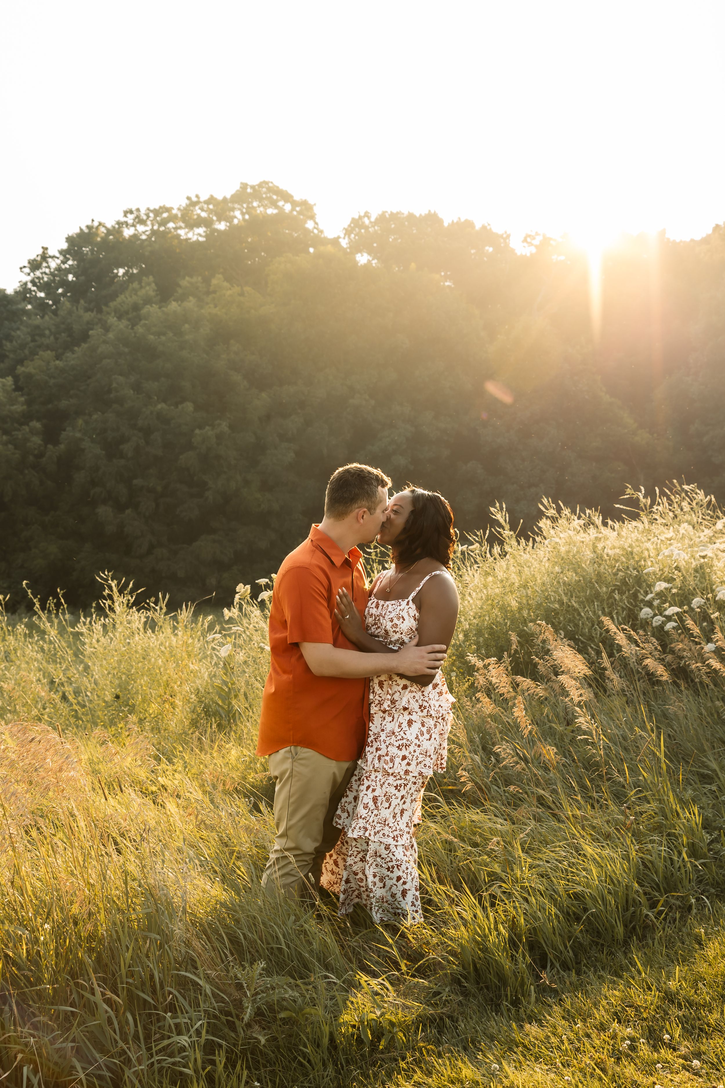 Couple sharing a romantic moment in a sunlit meadow, man in orange shirt embracing woman in floral dress at golden hour.