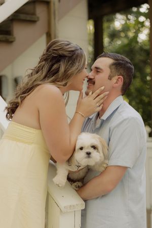 Couple sharing a romantic moment while holding a small white Shih Tzu dog on a white porch railing.