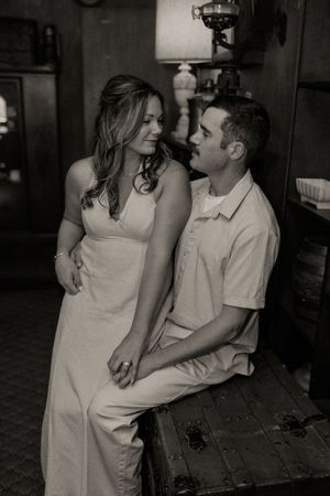 Black and white photo of a couple sharing a romantic moment, woman in white dress and man in polo shirt leaning on vintage trunk.