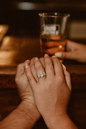 Close-up of two hands clasped together displaying a three-stone diamond engagement ring on a wooden surface.