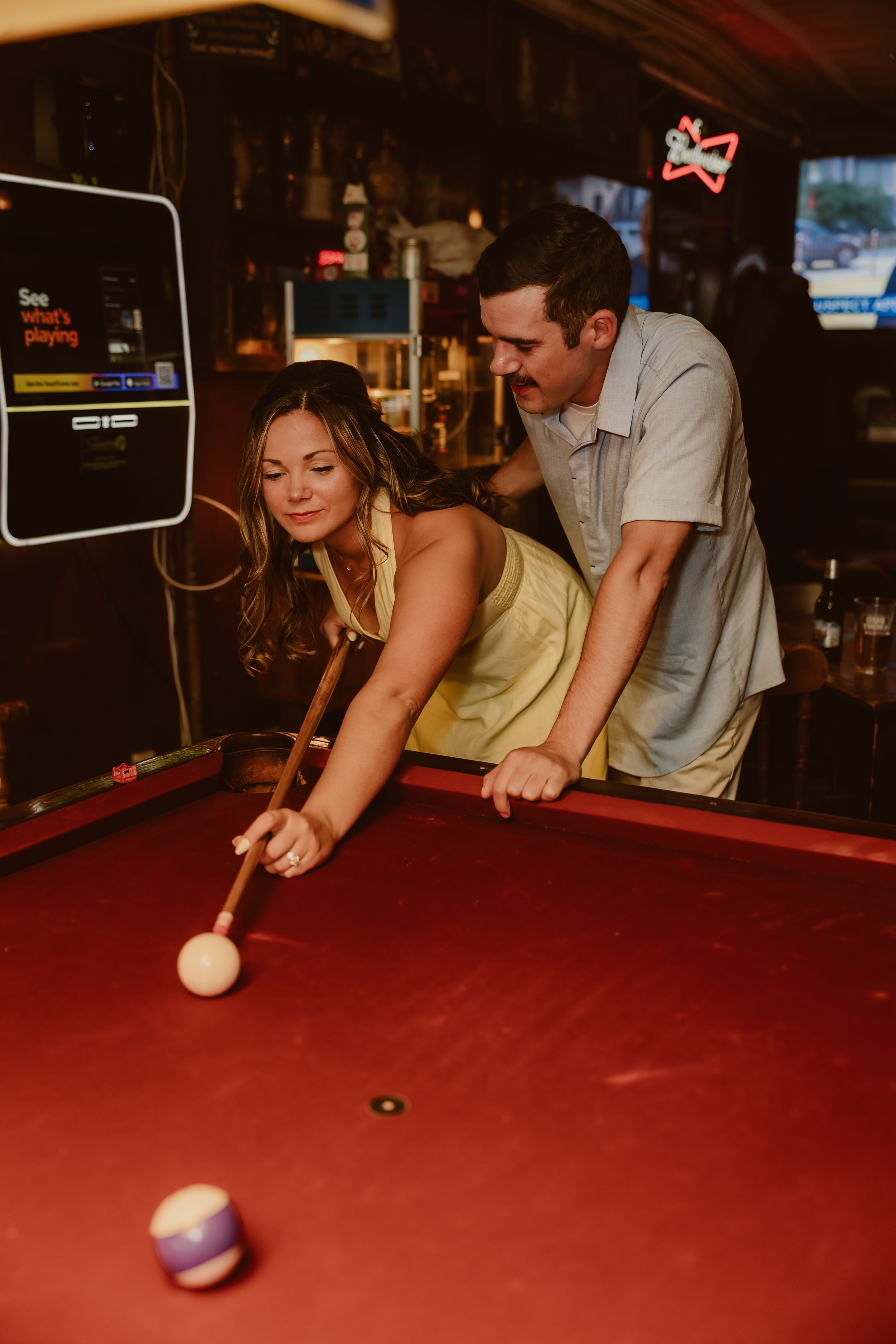Couple playing pool together at a bar, woman leaning over red billiard table taking a shot while man stands behind her.