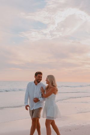 A couple walks hand in hand on a beach at sunset, dressed in white, with pastel skies and ocean waves behind them.