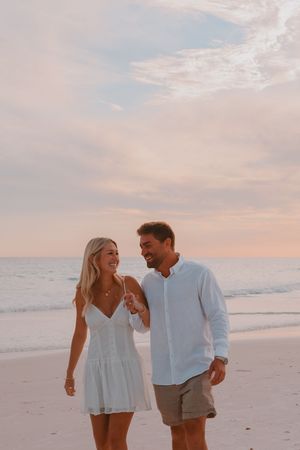 A smiling couple walks hand-in-hand on a sandy beach at sunset, wearing casual summer outfits.