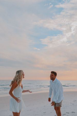Joyful couple laughing and dancing on a white sandy beach during a colorful sunset.