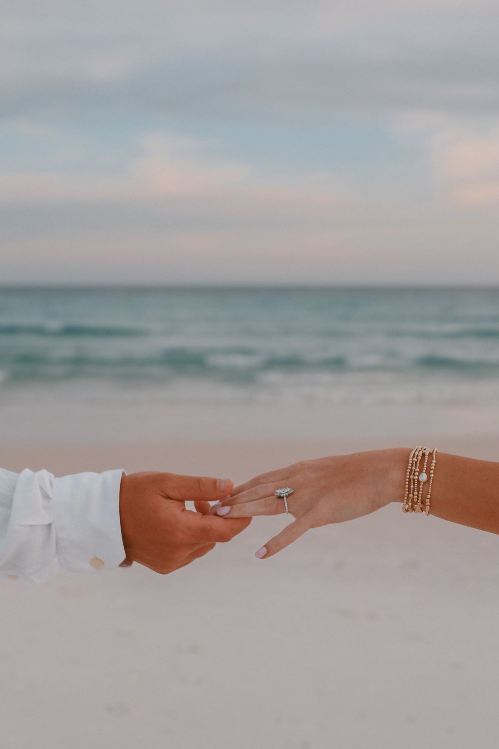 Close-up of hands on sandy beach, man placing diamond engagement ring on woman's finger.