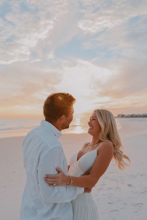 Couple embracing on a sandy beach at sunset, woman in white dress smiling, man in white shirt, pastel sky backdrop.