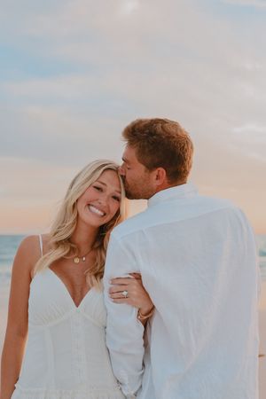 Man kissing smiling blonde woman on forehead during romantic beach engagement session at sunset.