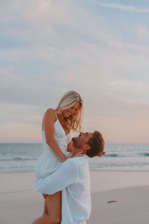 A couple laughing joyfully on a beach at sunset, man lifting woman in white dress near ocean waves.