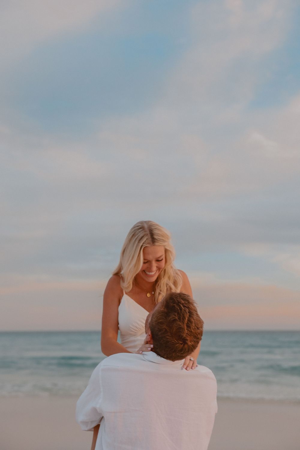 Couple sharing a romantic beach moment at sunset, woman in white dress smiling while man holds her close.