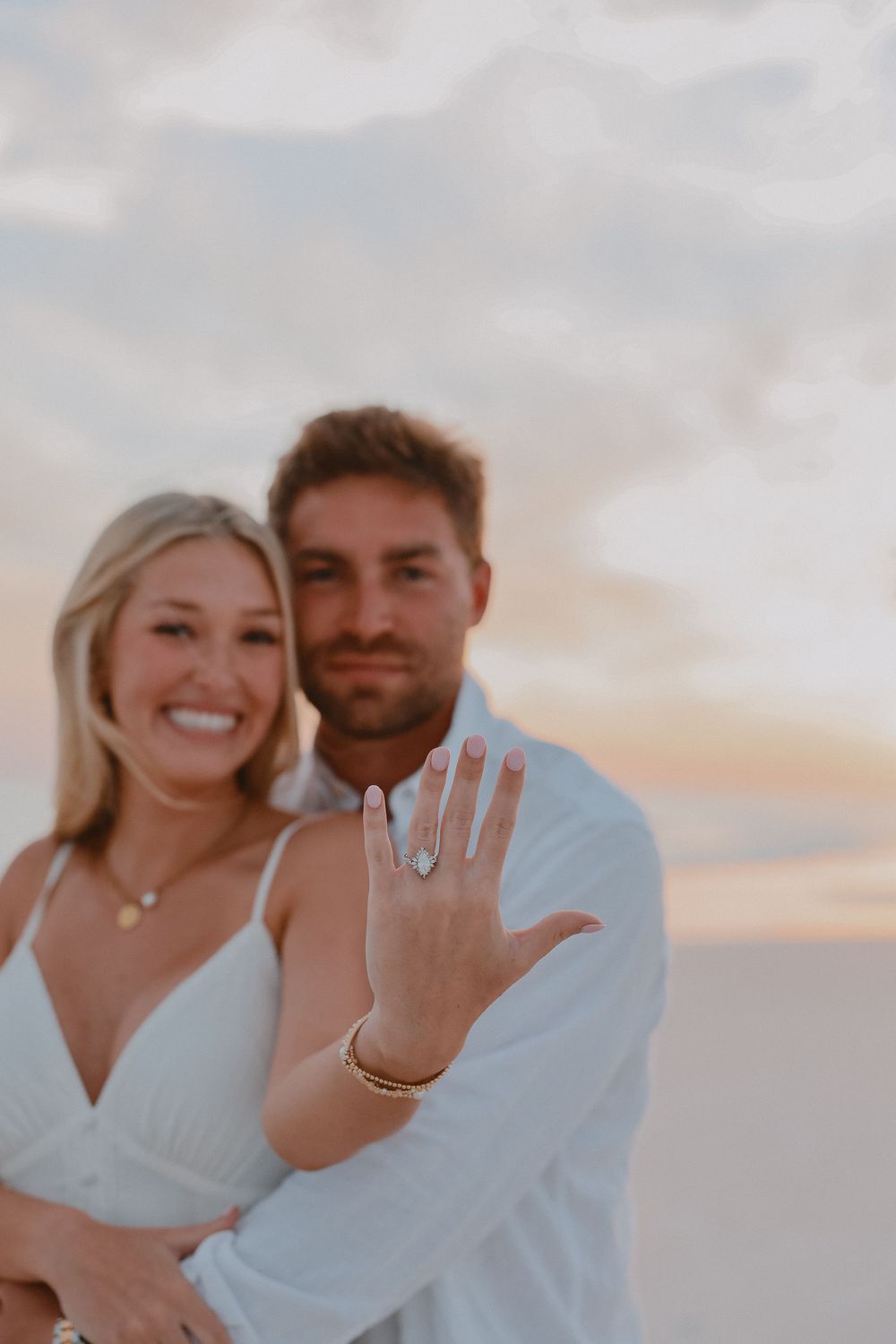 Newly engaged couple smiling on beach at sunset, woman showing off sparkling engagement ring.
