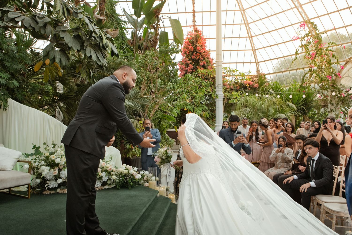 Bride and groom exchange vows in stunning glass greenhouse wedding venue filled with greenery.