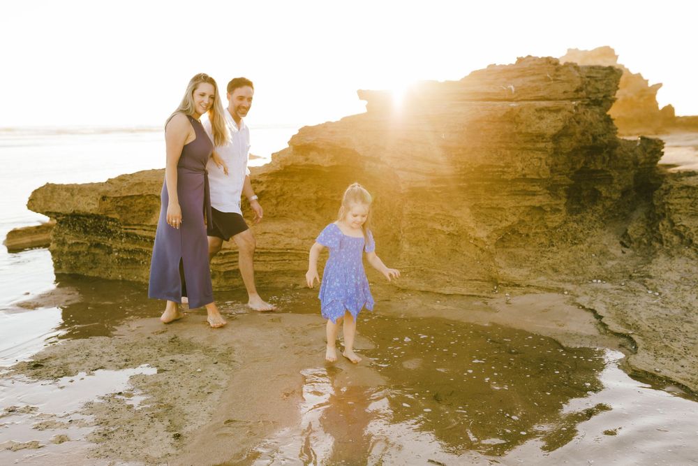 Family of three enjoying a sunny beach day near rocky formations, with a little girl in a blue dress playing in the water.