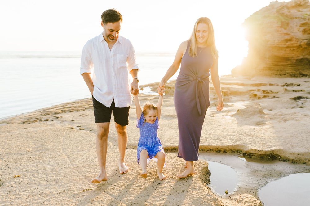 Family of three walking on rocky beach at sunset, parents holding toddler's hands in blue outfit.