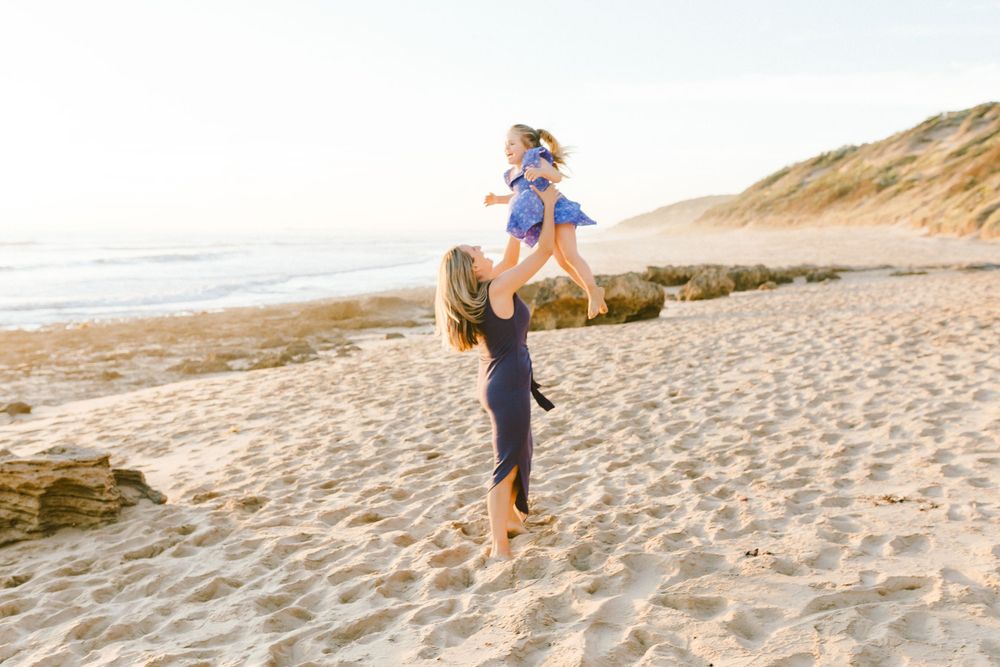 A mother joyfully lifts her young daughter in the air on a sunny sandy beach with ocean waves in the background.