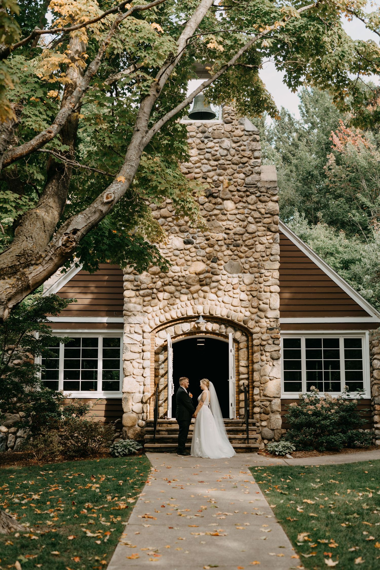 Bride and groom stand at entrance of rustic stone chapel surrounded by autumn trees with fallen leaves on pathway.