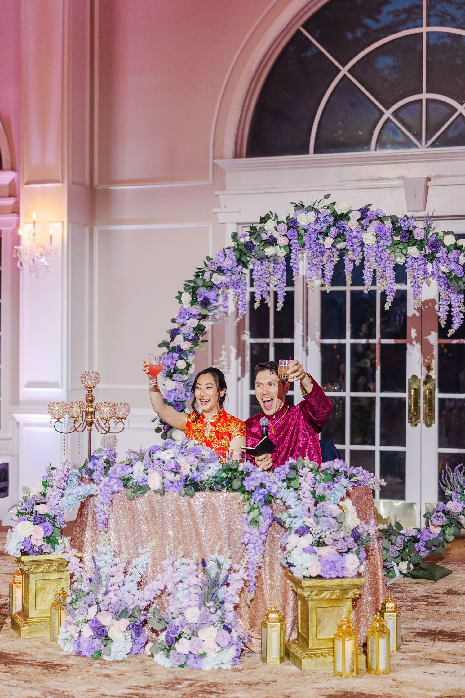 Couple in traditional Chinese wedding attire toasting at floral arch decorated with purple wisteria and hydrangeas in elegant ballroom.