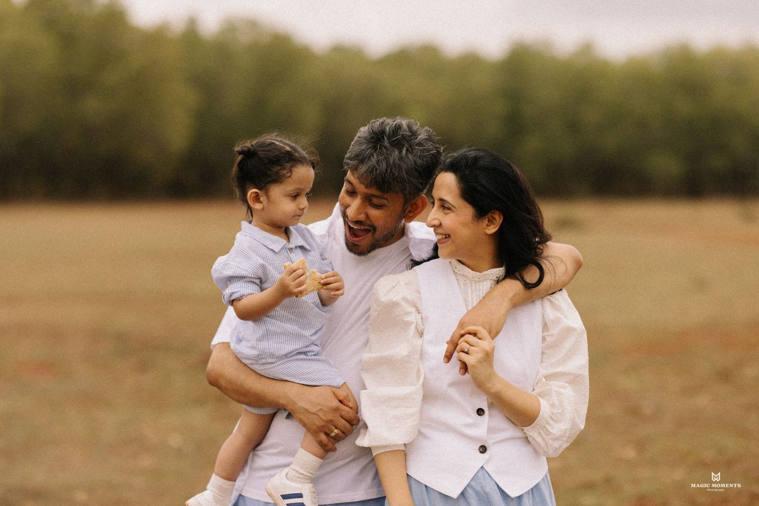 A smiling South Asian family of three poses together, with two women in red outfits flanking a man in a black shirt.