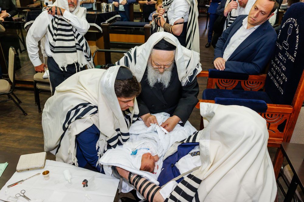 Jewish men in tallitot perform a bris circumcision ceremony, gathered around a newborn baby in a synagogue.