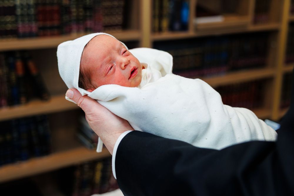 Newborn baby wrapped in white blanket and cap, gently cradled in adult hands near a bookshelf.