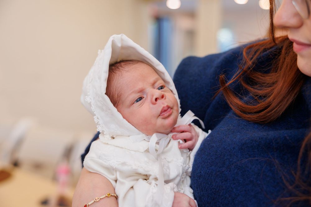 Newborn baby in white bonnet cradled in arms of woman in blue sweater.