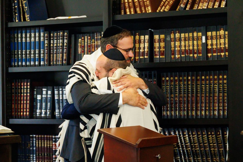 Two men in Jewish prayer shawls share an emotional embrace near a podium in front of a bookshelf of religious texts.