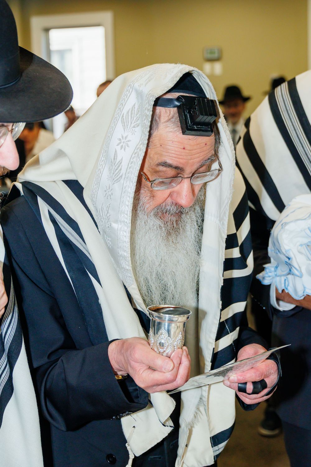 Elderly Orthodox Jewish rabbi wearing tallit and tefillin holds kiddush cup during religious ceremony.