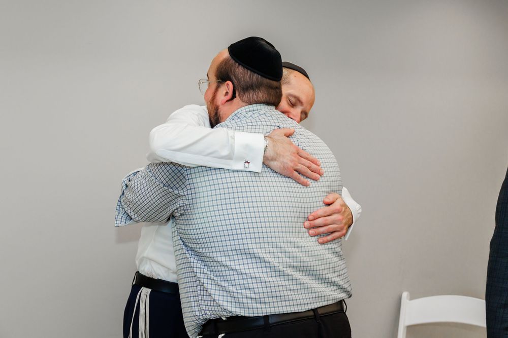Two men wearing kippot share a warm embrace in a bright indoor setting.