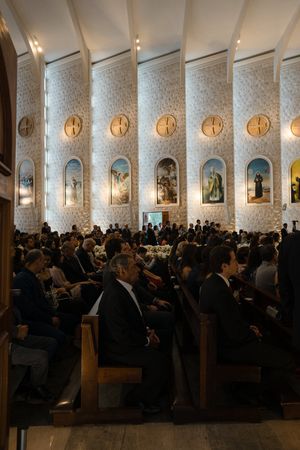 A crowded church congregation sits in wooden pews during a service with decorative wall panels and lighting fixtures.