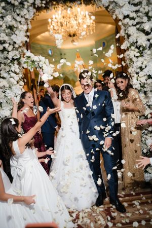 Wedding party celebrates with flower petals being tossed in the air under an elegant chandelier at an indoor venue.