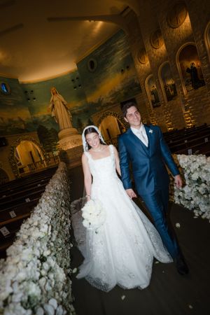 A wedding couple walks through an ornate ballroom decorated with white floral arrangements and dramatic lighting.