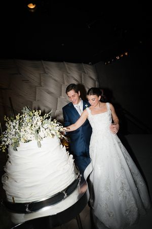 A couple in wedding attire cuts into a tall white wedding cake with floral decorations in a dimly lit reception.