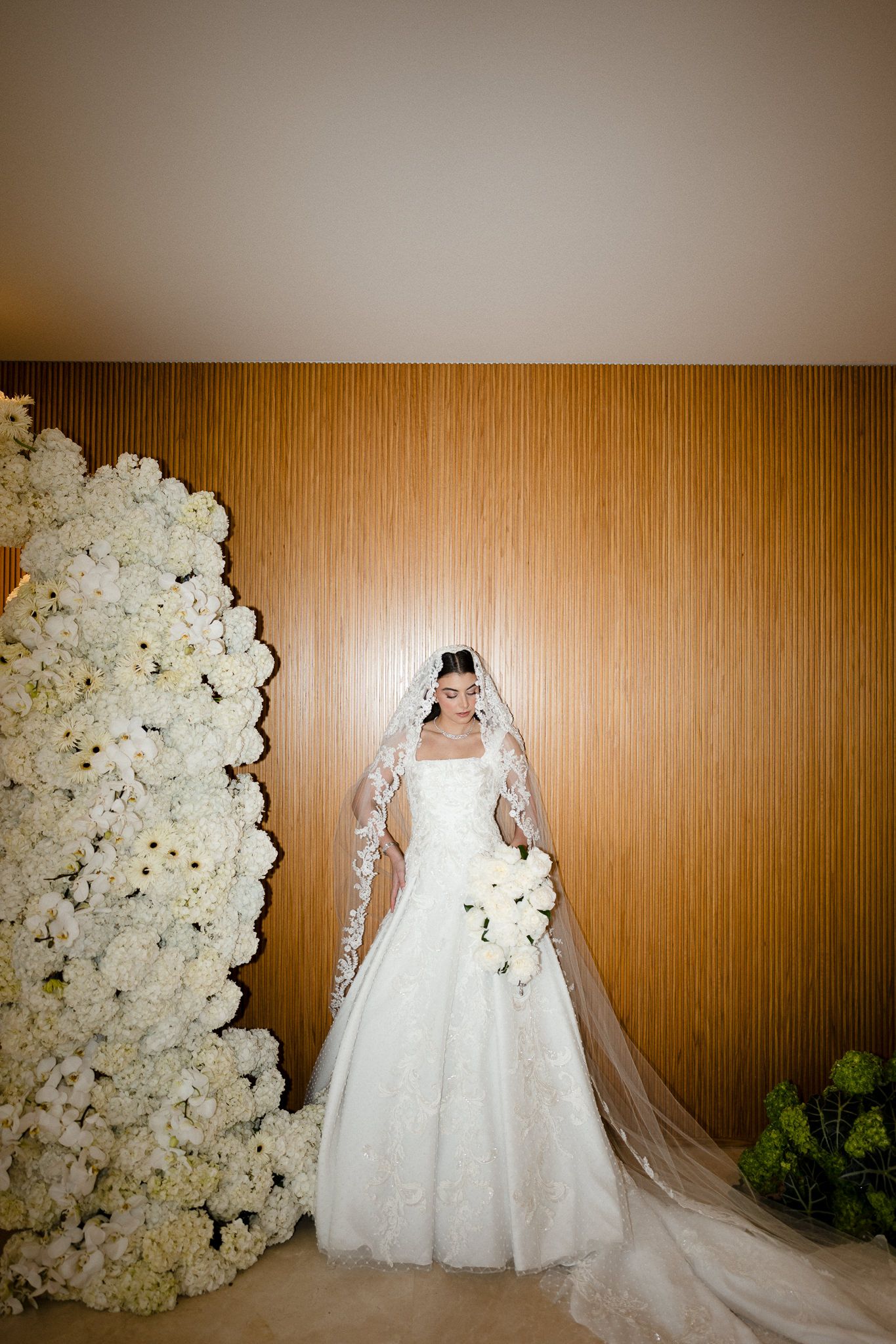 Wedding dress displayed against wood paneling with cascading white floral wall installation creating an elegant backdrop.