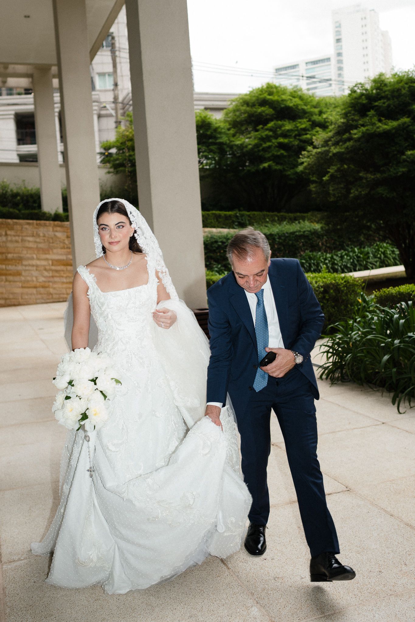 A bride in a white wedding dress sits on a concrete ledge while a man in a navy suit kneels beside her.