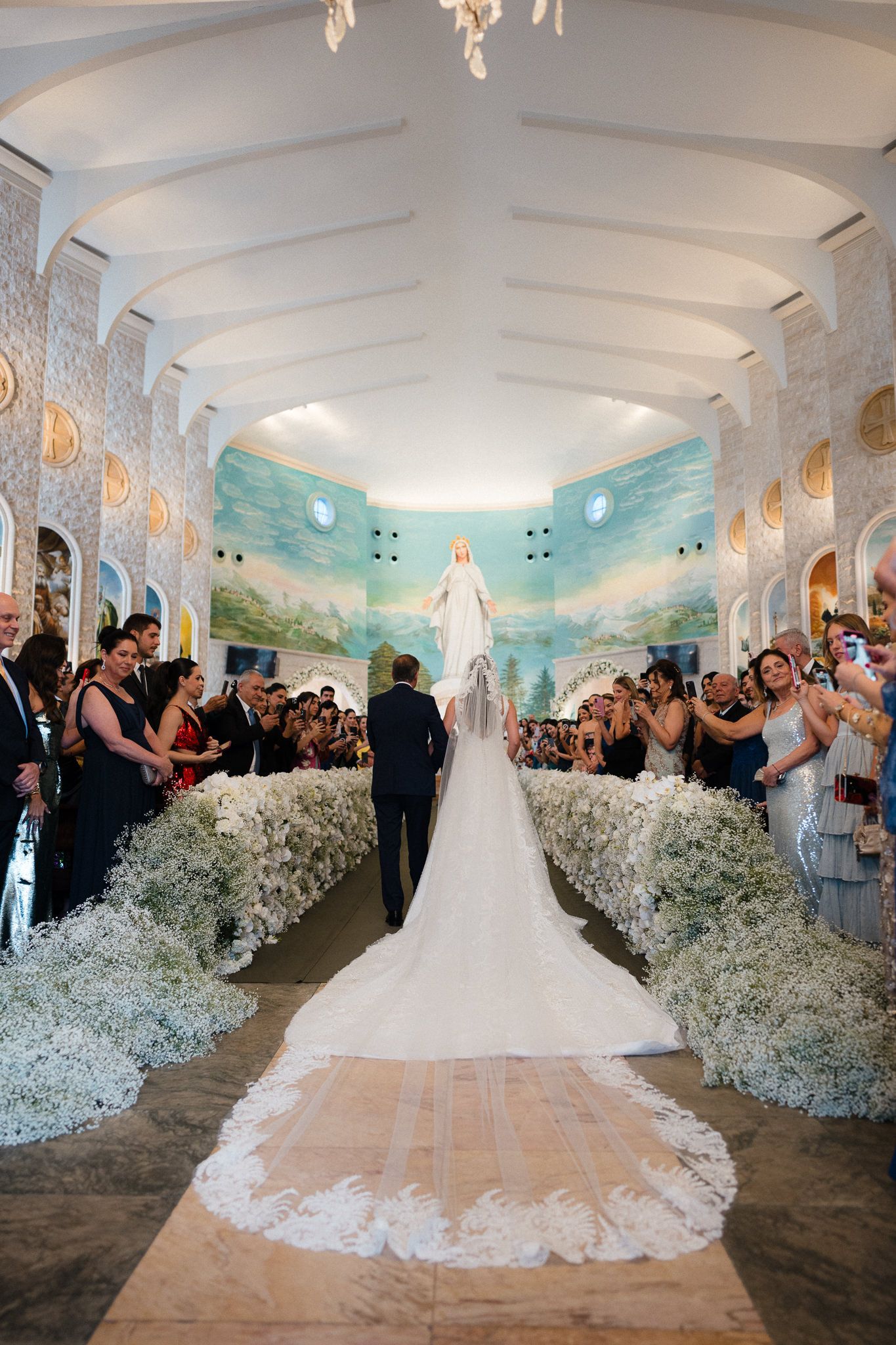Elegant wedding ceremony in a white room with painted murals and baby's breath floral arrangements lining the aisle.