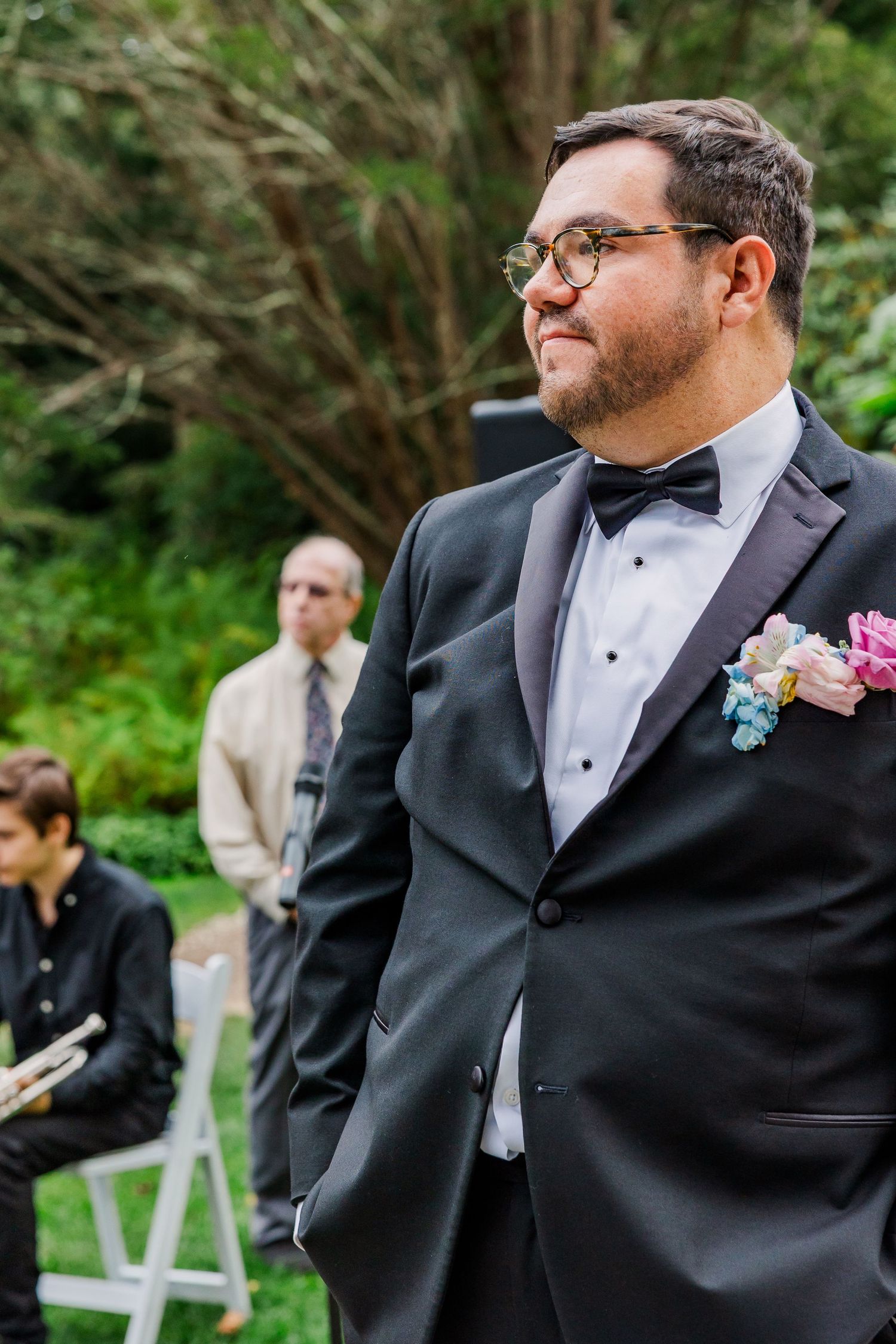 A groom in a black tuxedo and bow tie with a floral boutonniere stands during an outdoor wedding ceremony.