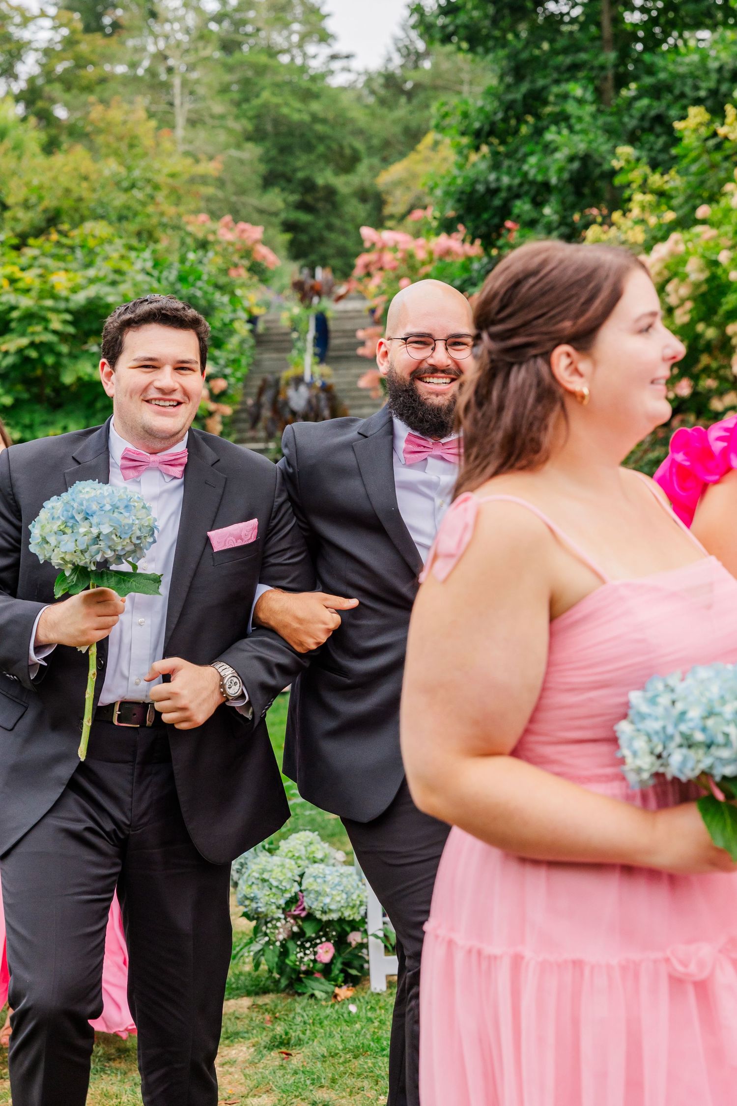 Wedding party members in pink dresses and black suits share candid laughing moments during outdoor photo session.