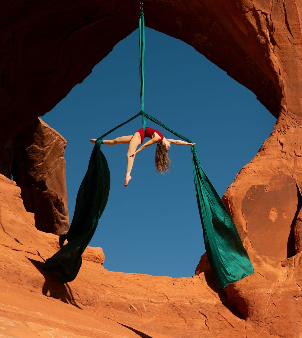 Aerialist suspended on silks in a Utah red rock arch; bold aerial arts photography combining clean lines, color, and natural texture.