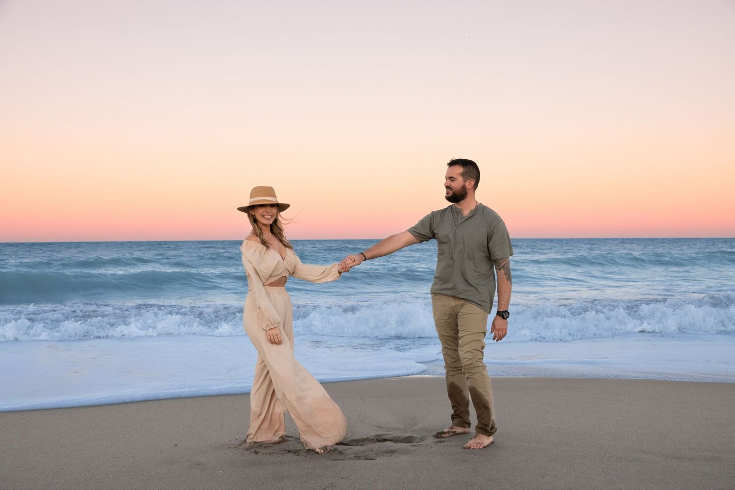 Couple holding hands on beach at golden sunset, woman in flowing dress and hat.