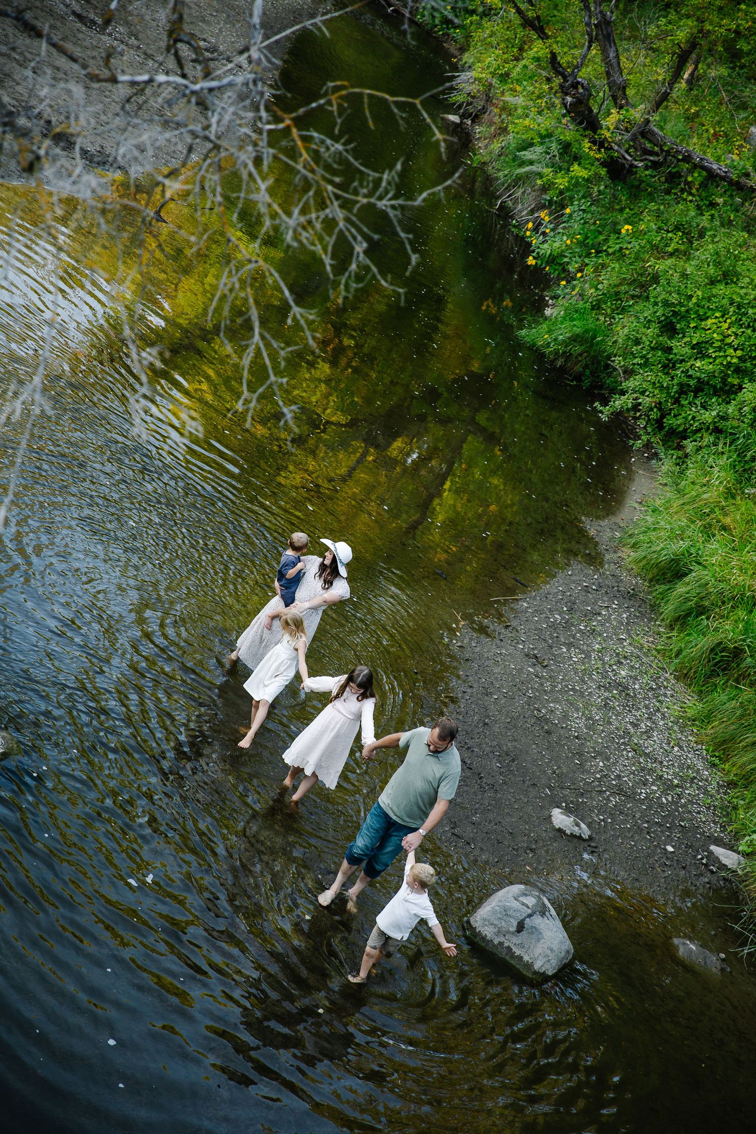 A couple wades through shallow water near a rocky riverbank surrounded by lush greenery.