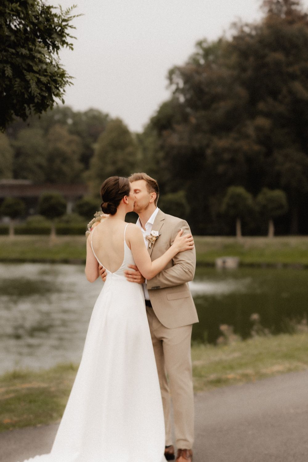 A couple shares a romantic moment by a lake in a park setting during what appears to be a wedding photoshoot.