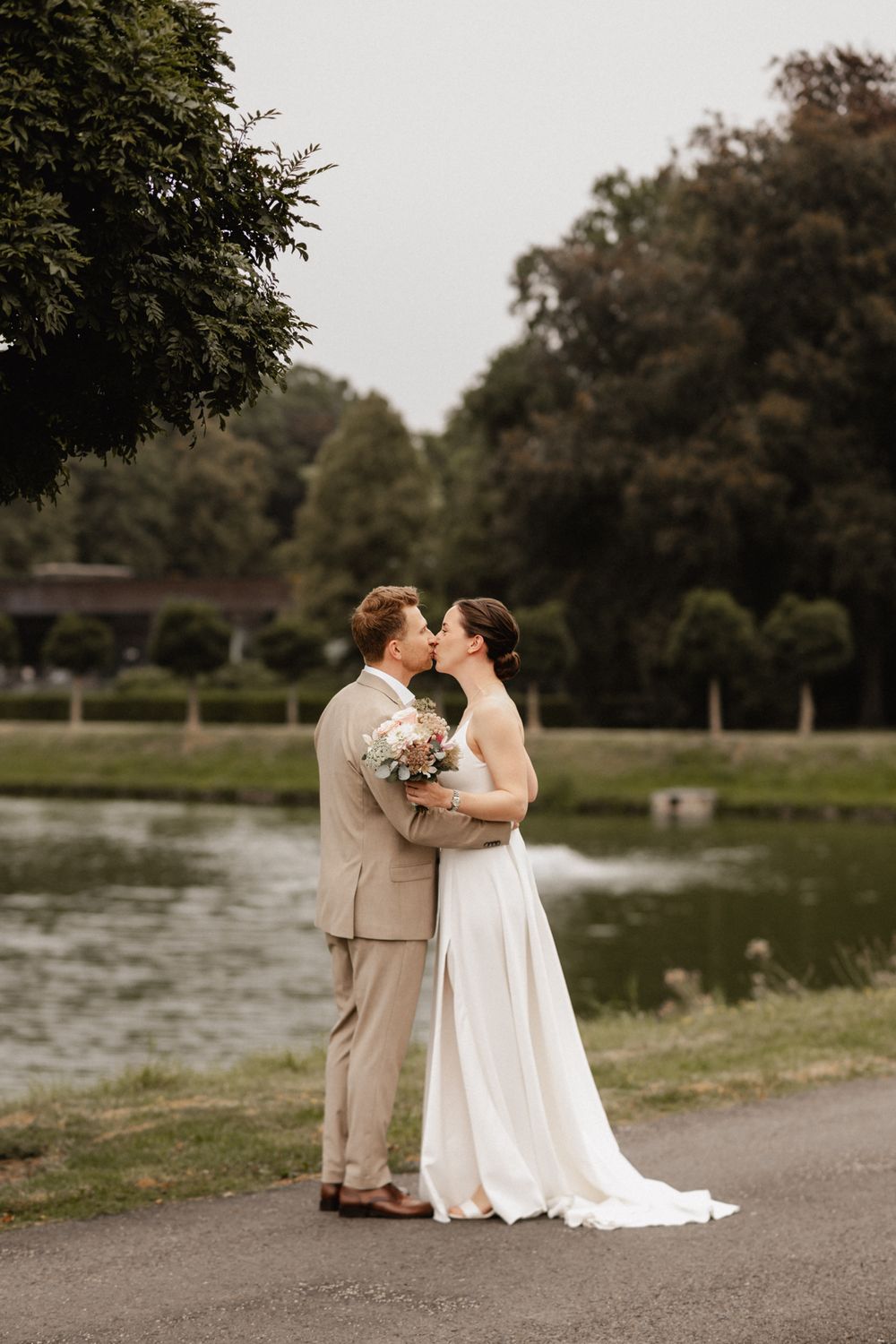 A couple in wedding attire share a romantic moment by a lake, with the groom in beige and bride in white gown.