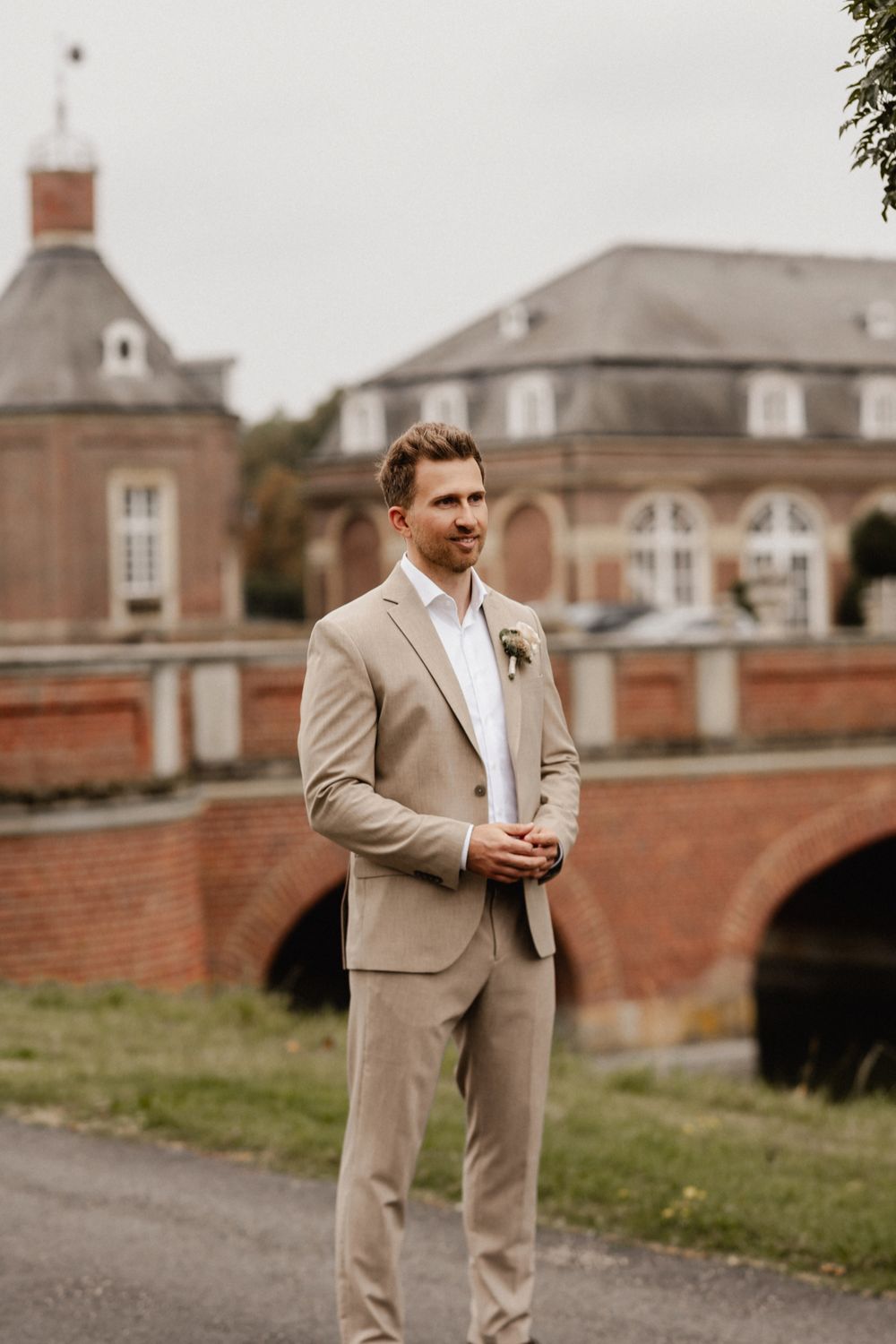 A groom in a tan suit stands outside a historic brick building with architectural details.