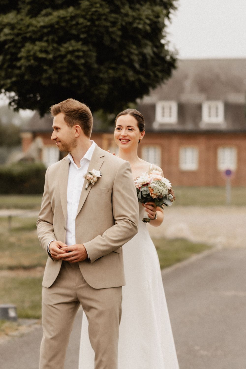 A couple shares a joyful moment in their wedding attire, with the bride in white and groom in beige suit outdoors.